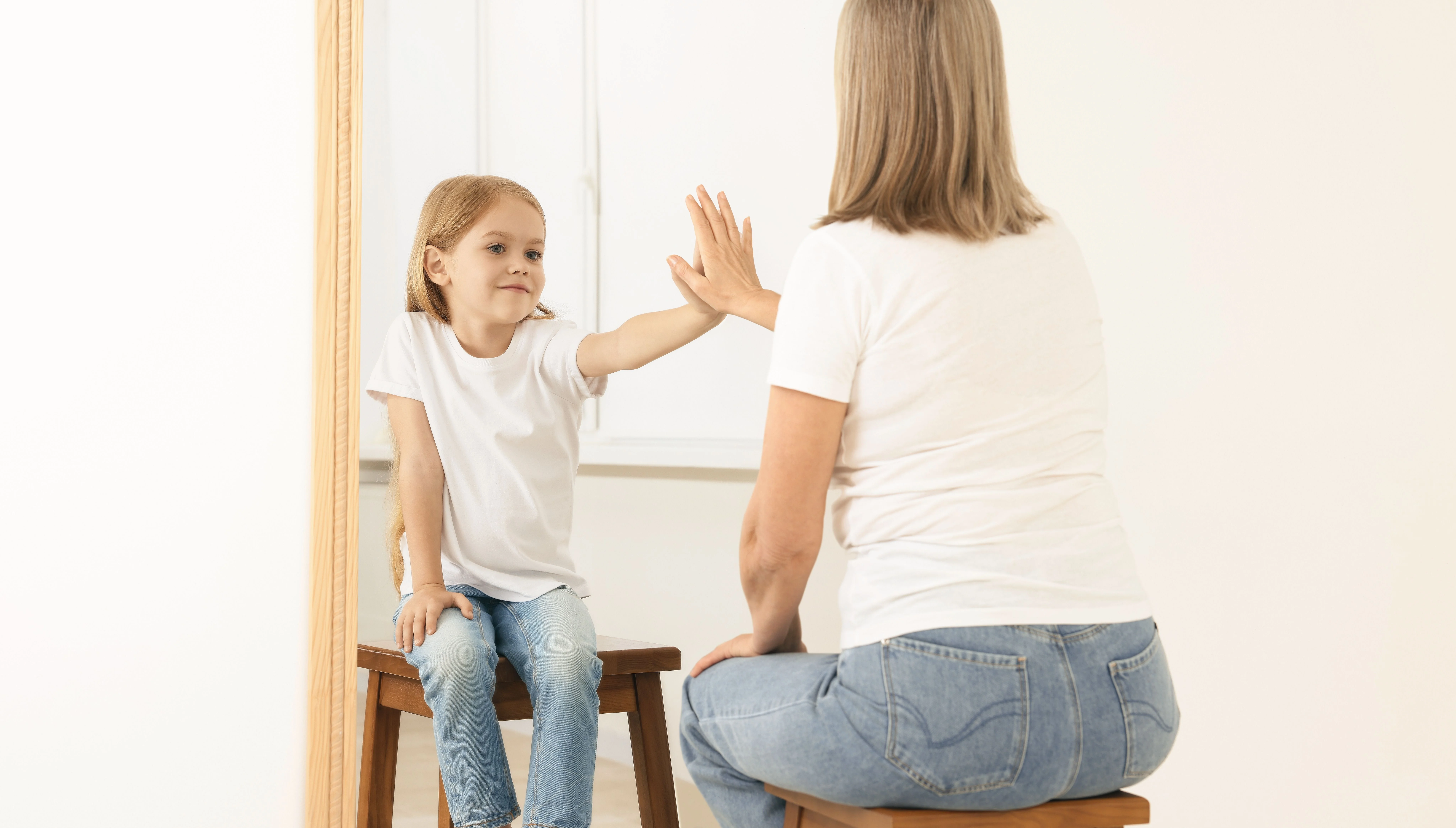 Una bambina e una donna sedute su sgabelli, si allungano l'una verso l'altra attraverso uno specchio in un momento gioioso