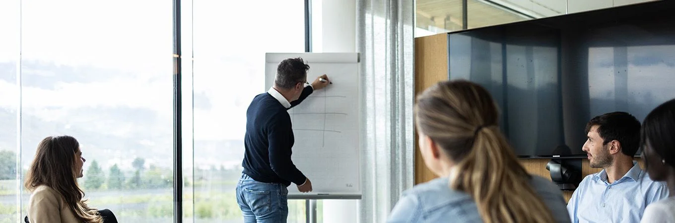 Ein Mann skizziert eine Strategie auf einem Flipchart, während Kollegen aufmerksam in einem hellen Besprechungsraum zuhören