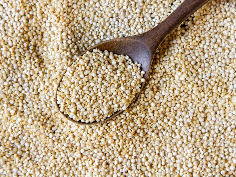 A wooden spoon holds a small mound of quinoa atop a larger pile of scattered quinoa grains
