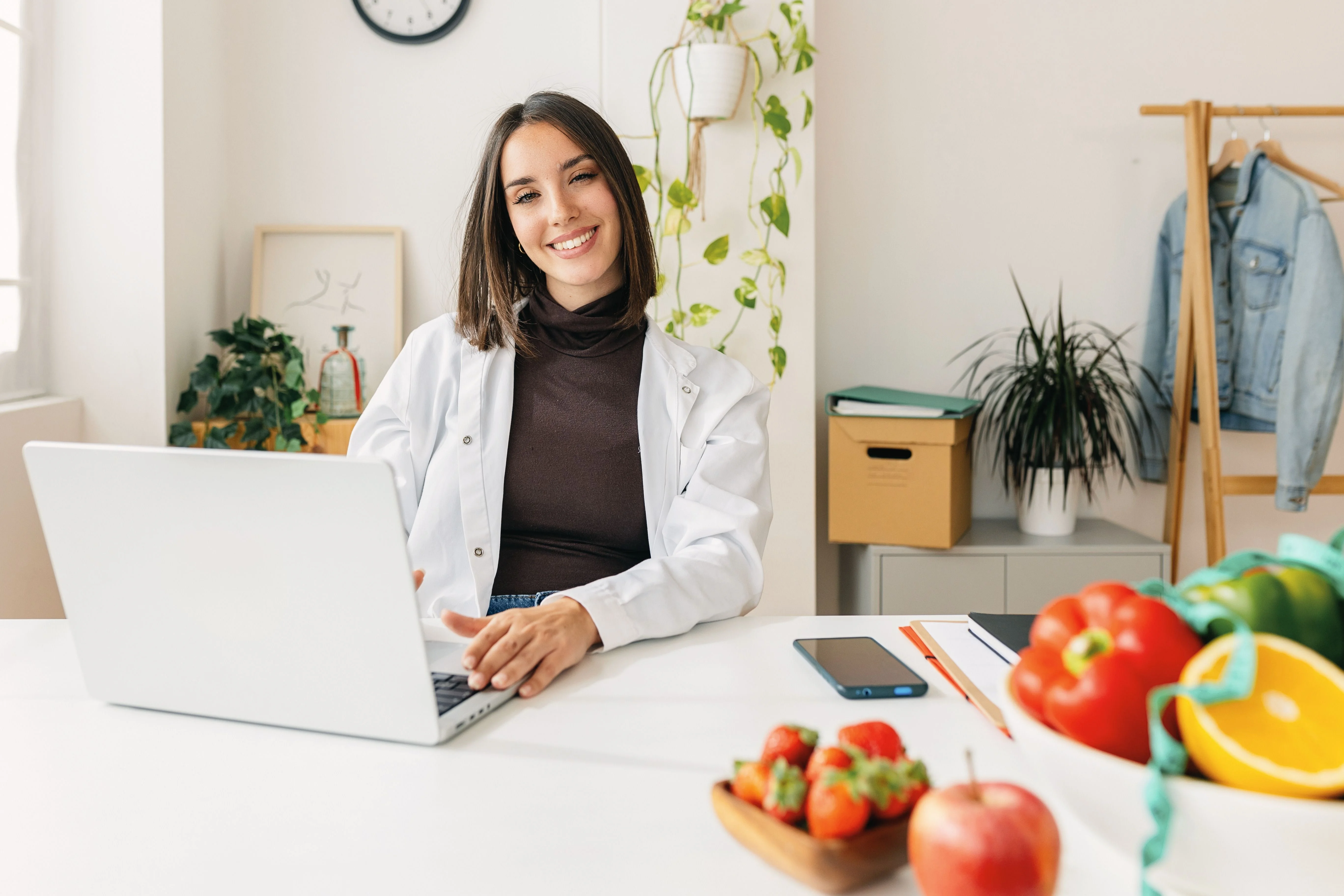 Smiling woman in a white lab coat sits at a desk with a laptop, surrounded by fresh fruits and greenery