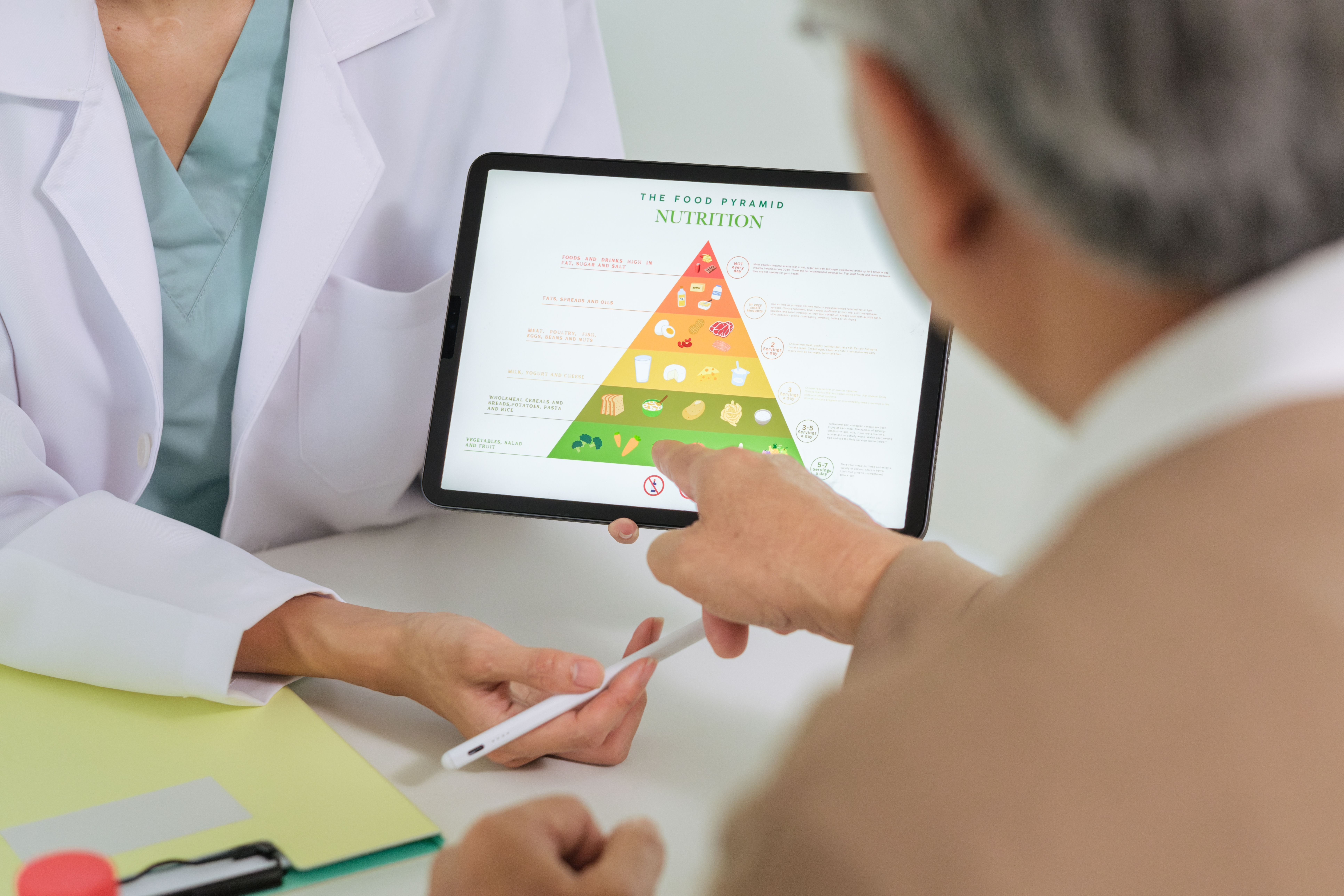 A healthcare professional points to a digital food pyramid on a tablet while discussing nutrition with a senior patient