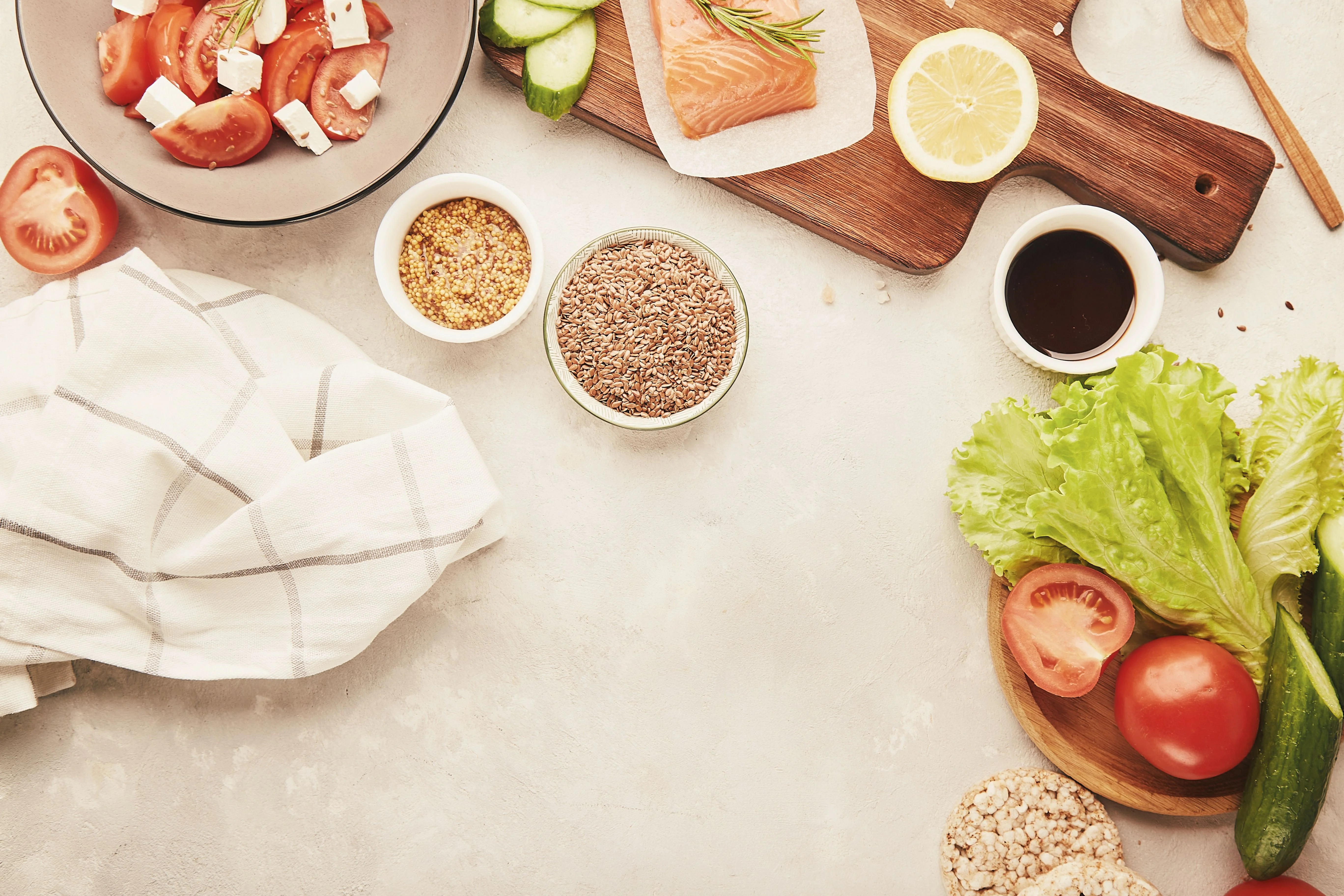 A spread of fresh ingredients, including tomatoes, cucumbers, lettuce, salmon, and mustard, on a light surface