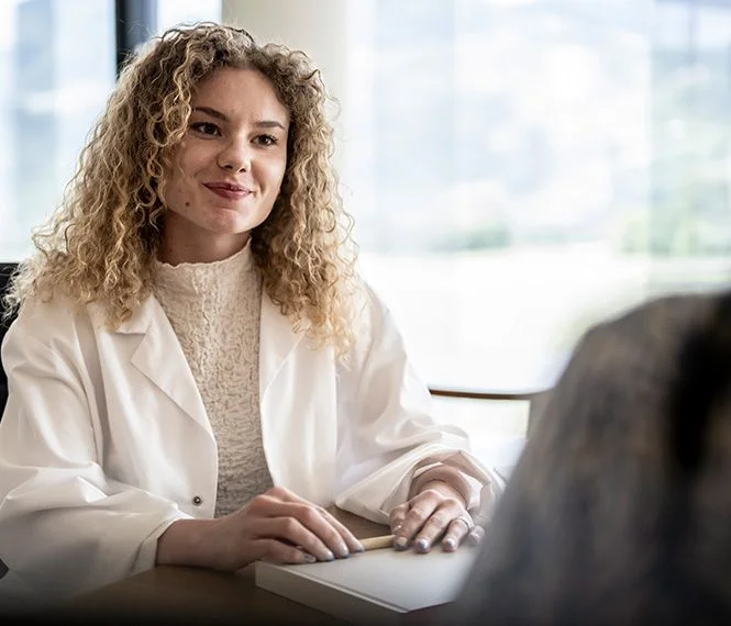 A smiling woman with curly hair in a white lab coat engages in conversation with a person out of view