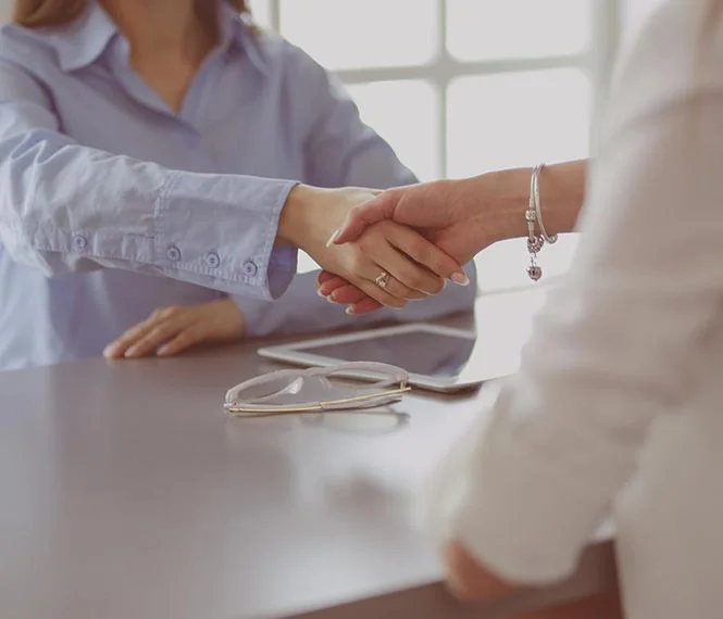Deux personnes se serrent la main au-dessus d'une table, avec des lunettes et une tablette à proximité, dans un bureau lumineux