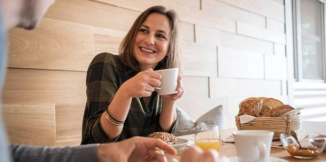 A woman in a checkered shirt smiles while holding a coffee cup at a cozy breakfast table with pastries