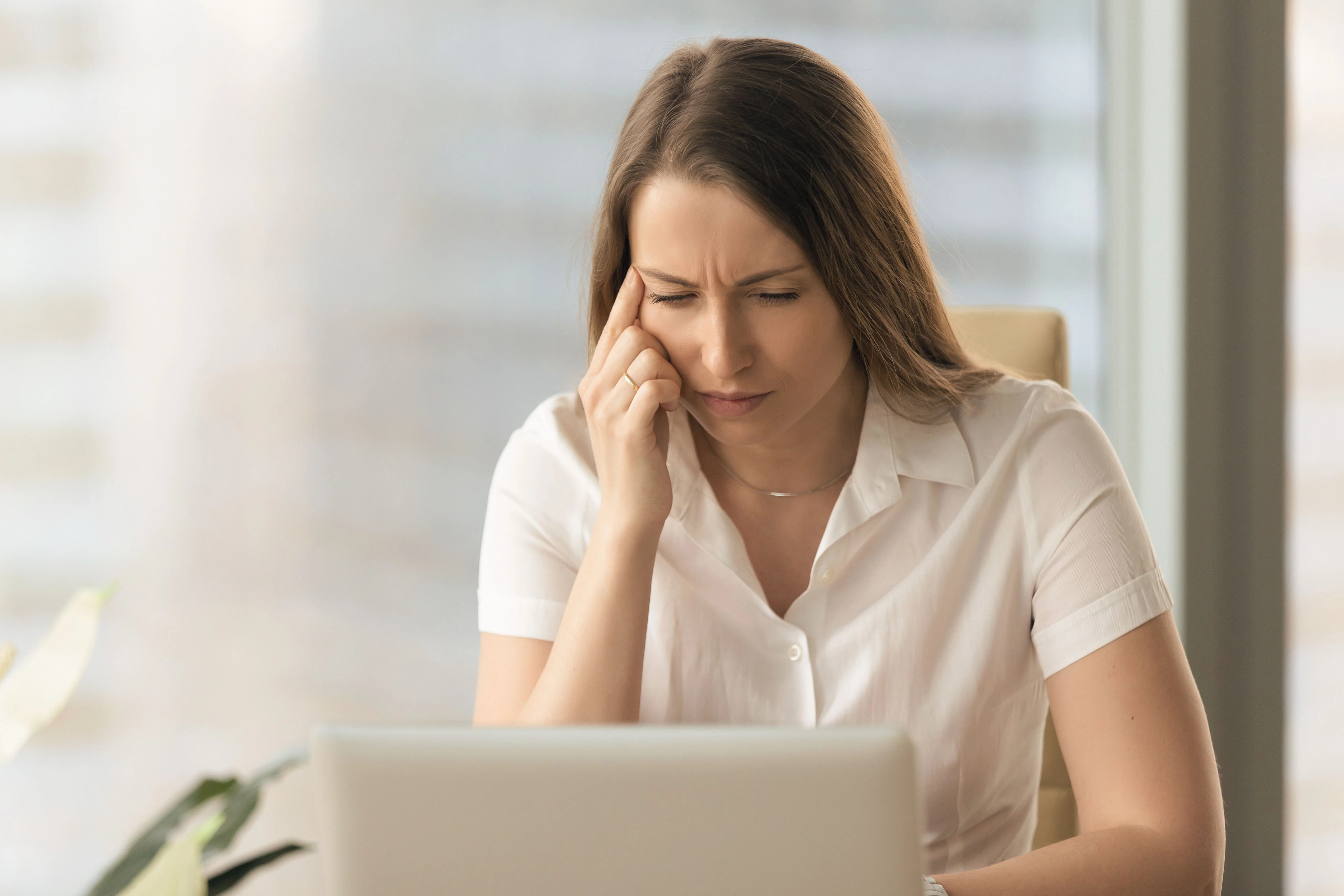 A woman in a white blouse looks stressed while working on her laptop in a modern office environment