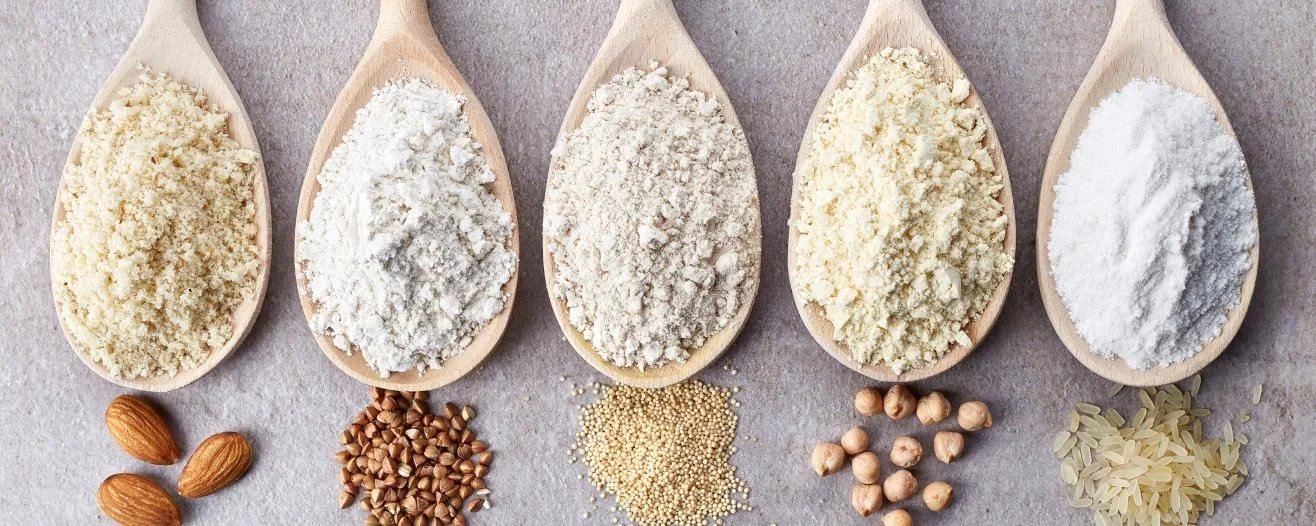 Cinque cucchiai di legno contenenti farine e polveri diverse accanto a mandorle e semi su una superficie di pietra